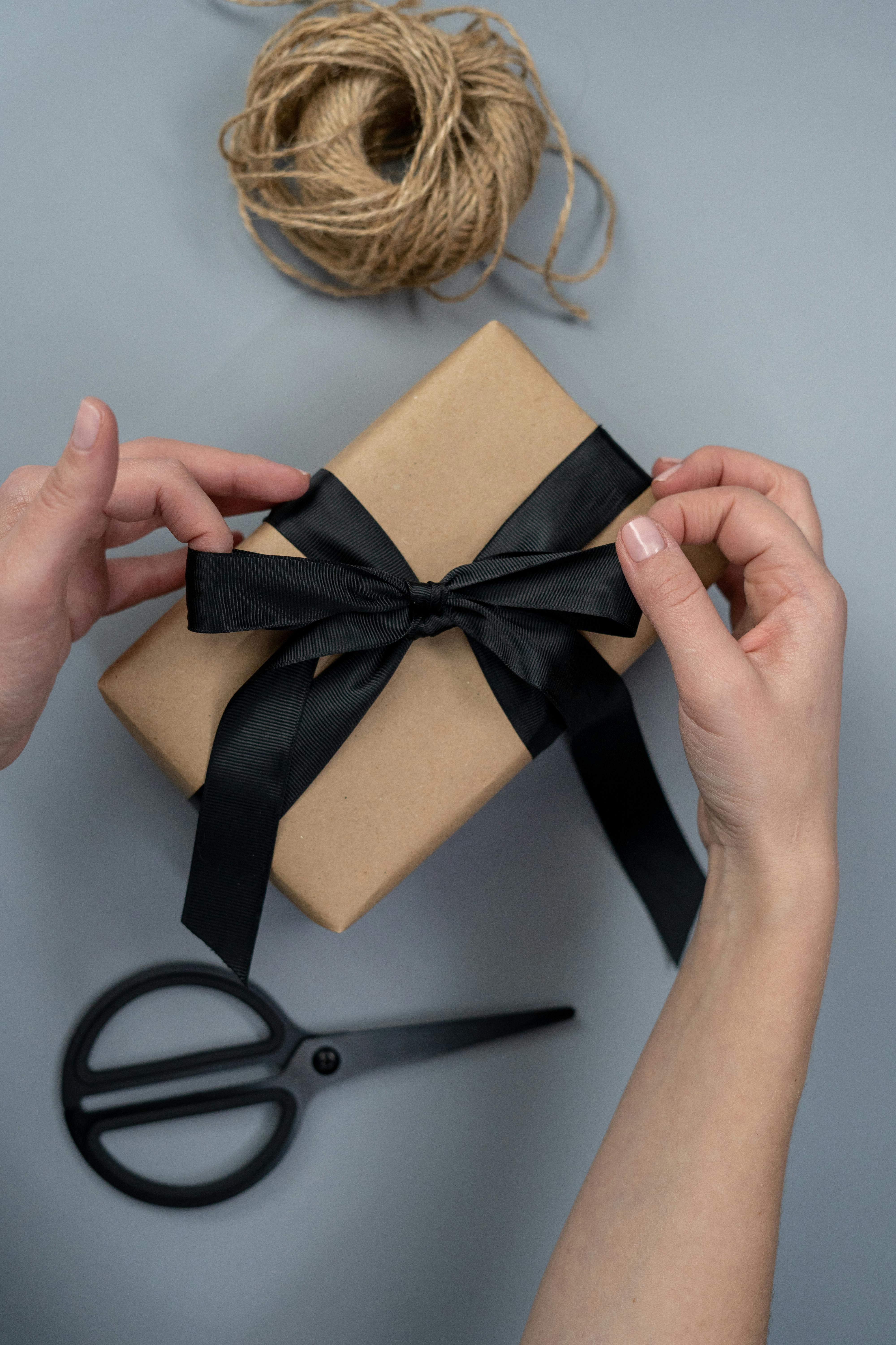 Hände binden eine schwarze Schleife um ein Geschenk in Packpapier, passend zu Geschenkideen, Buchgeschenken, Schreibwaren und liebevoll verpackten Produkten
